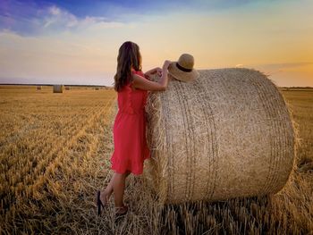 Rear view of woman in red dress standing near a hay bale at sunset