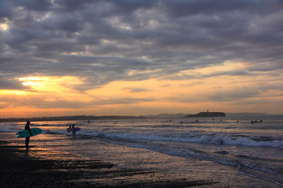 People with surfboard at sea shore against sky during sunset