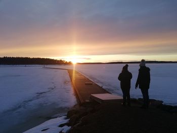 People standing on snow covered land against sky during sunset