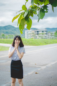 Portrait of young woman with hands covering mouth standing on road against cloudy sky