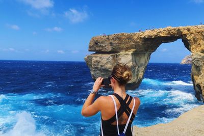 Rear view of woman by rock formation photographing sea