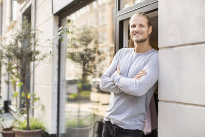 Portrait of smiling owner leaning on wall while standing outside clothing store