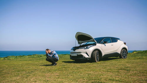 Frustrated man crouching against broken down car at beach