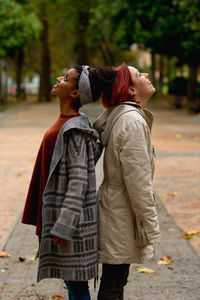 Side view of joyful content stylish multiethnic girl friends enjoying and looking up standing back to back in park