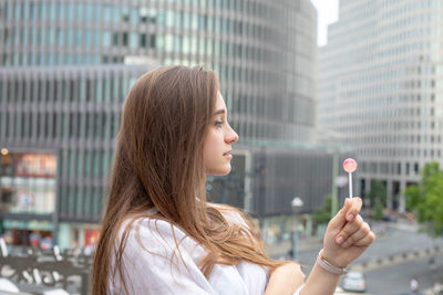 Side view of young woman holding lollipop by buildings in city