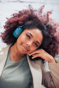 Glad black businesswoman in suit with curly hair touching face and looking at camera while listening to music on street in daytime