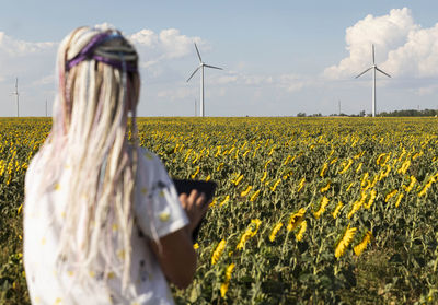 Young woman with tablet in field with sunflowers, wind turbines, green energy production, eco-energy