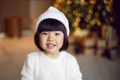 Beautiful korean child girl in in a white knitted sweater and hat stands at the christmas tree 