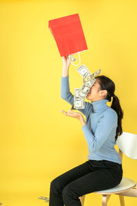 Side view of young woman sitting against yellow background