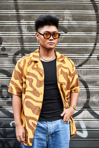 Stylish serious young non binary male in bright shirt and sunglasses with arms crossed looking away while standing against metal wall