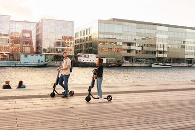 Full length of couple riding electric push scooters on promenade by river in city