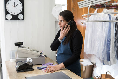 Mature female dry cleaner talking on smart phone while standing at checkout in laundromat