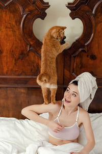 Young woman with a red cat on his shoulder, towel on his head sits in an old bed, smiling.