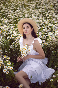 Beautiful young brunette woman in a hat and a white dress standing on a chamomile field at sunset