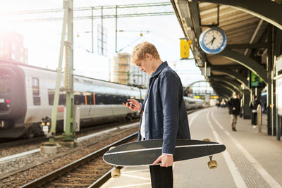 Side view of young man using mobile phone while holding skateboard on railroad station platform