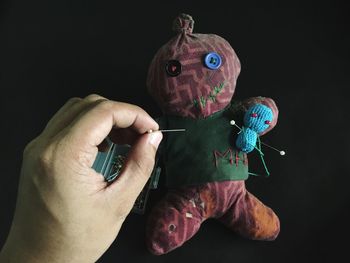 Cropped hand of man putting straight pin in voodoo doll over black background