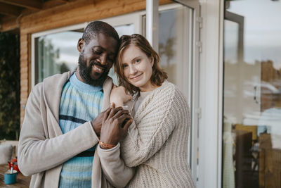 Portrait of smiling young woman embracing man while standing on porch