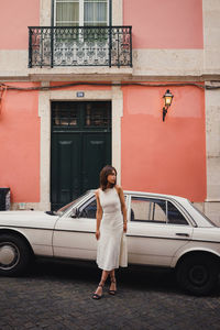 Full body positive female in dress walking near white old timer automobile on street with shabby building in city