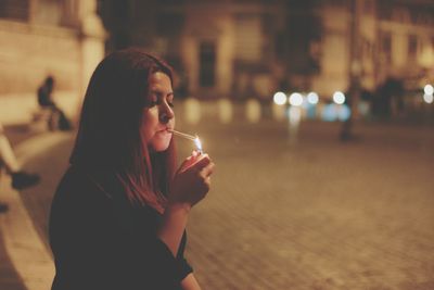 Woman igniting cigarette by street in city at night