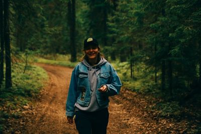 Portrait of smiling woman wearing cap walking on dirt road amidst forest