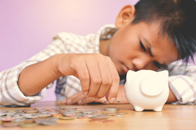Close-up of boy putting coin in piggy bank at table against wall