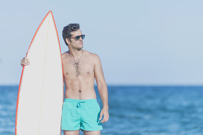 Shirtless man holding surfboard while standing against sea