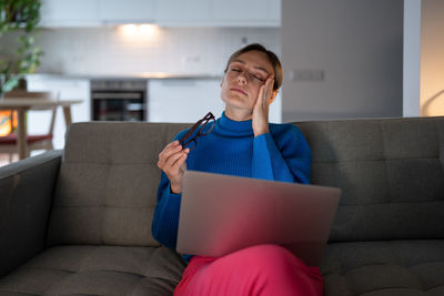 Lonely woman sits on comfortable sofa with exhausted expression and procrastinates with work