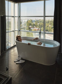 Woman taking bath in bathtub against window at luxury hotel room