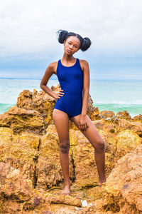 Portrait of teenage girl in swimwear standing on rock at beach against sky