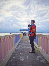 Rear view of man standing on railing against sky