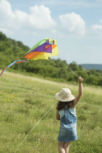 Rear view of girl holding kite while walking on field against sky