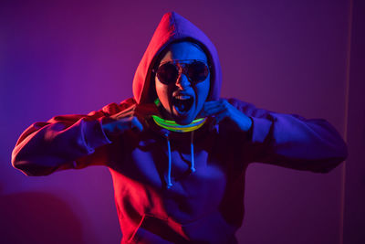 Cyberpunk woman in a hooded hoodie and sunglasses dances against a wall with neon sticks hanging 