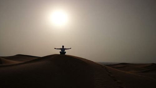 Man sitting on sand dune with arms outstretched against clear sky