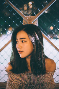 Thoughtful beautiful woman looking away against fence at night