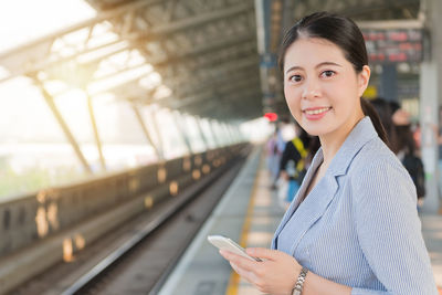 Portrait of smiling woman holding smart phone while standing at railroad station