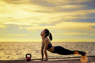 Side view of young woman exercising at beach against sky during sunset