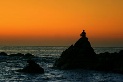 Silhouette man siting on rock over sea against orange sky