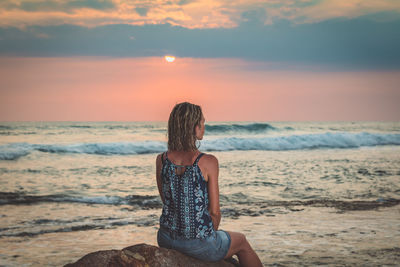 Young woman sitting on beach against sky during sunset