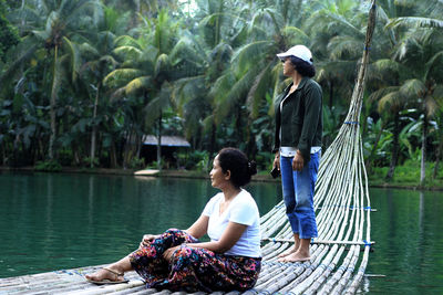 Rear view of friends sitting by lake against trees