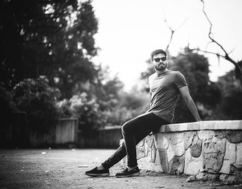 Full length of young man looking away while sitting on retaining wall