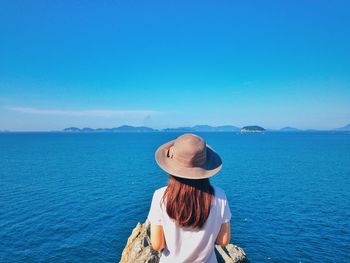 Rear view of woman looking at sea while standing against clear blue sky during sunny day