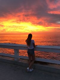 Rear view of woman standing by railing at beach against cloudy sky during sunset