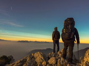 Rear view of hikers standing on cliff against sky during sunset