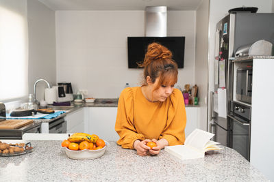Caucasian woman dressed in orange t-shirt sitting at the kitchen table reading a book