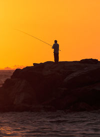 Man fishing while standing on rock by sea against orange sky