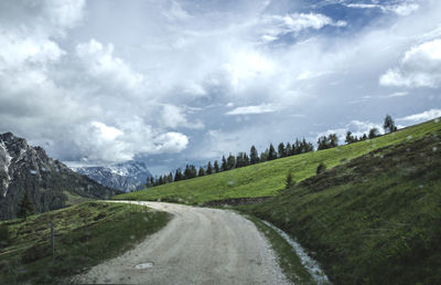 Dirt road amidst green hill against cloudy sky