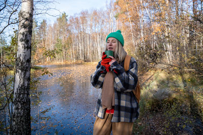 Freezing middle aged tourist woman hiker drinking hot tea from cup enjoying nature forest lake.