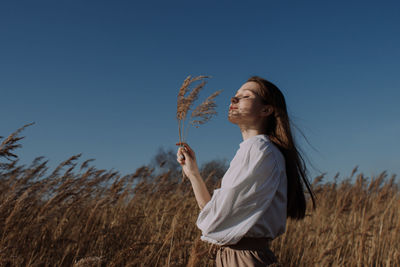 Young woman in white blouse standing in field and holding branch of dry pampas grass in front of sky
