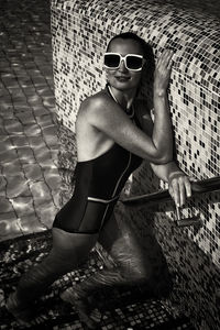A vintage photo of a woman in a black swimsuit and white sunglasses sunbathing by the pool wall