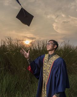 Young man wearing graduation gown while throwing cap against sky during sunset